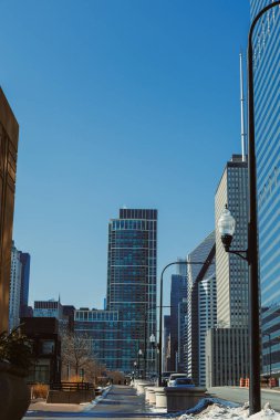 Gündüz vakti parlak mavi gökyüzüne uzanan modern binaların görüntüsü. Araçlar yol boyunca yürür, sokak lambaları da yürüyüş yolunda..