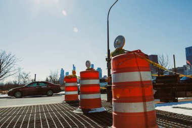 İşçiler, araçlar geçerken cadde boyunca parlak turuncu inşaat bariyerleri yerleştiriyorlar. Açık mavi gökyüzünün altında arka planda yüksek binalar yükseliyor.