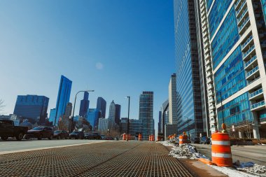 Şikago şehir merkezindeki caddede inşaat bariyerleri var ve yakınlarda arabalar var. İnsanlar yoğun bölgede hareket ettikçe, yüksek binalar mavi gökyüzüne doğru yükseliyor..