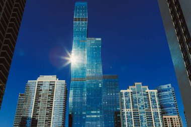 Parlak güneş ışığı hareketli bir şehirde uzun cam binalardan yansıyor. Sahne, mavi gökyüzü ve bölgeyi çevreleyen modern mimariyle açık bir günü yakalıyor..