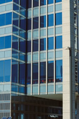 İnsanlar büyük pencereli yüksek bir binanın içindeki masalarda oturuyorlar. Güneş ışığı camdan yansıyor, kentsel bir alanda canlı bir sahne gösteriyor..