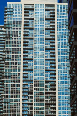 Cam cepheli uzun binalar şehirde güneş ışığını yakalıyor. Yapılar birbirine yakın duruyor, açık bir günde kentsel yaşamın yoğun ortamını gösteriyorlar..