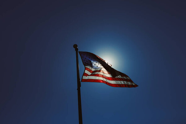 The US flag is raised on a pole and contrasts with the sun shining behind it. The clear sky gives a bright backdrop to the flag. This scene shows a moment of national pride.