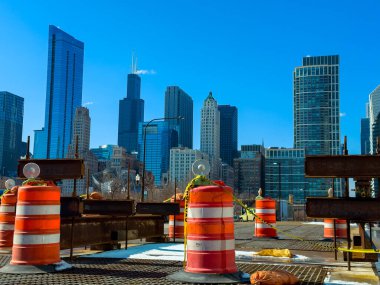 Şikago 'da inşaat çalışmaları yapılıyor ve turuncu koniler bölgeyi işaretliyor. Gökdelenler arka planda açık mavi gökyüzünün altında. Gündüz gözüyle kalabalık bir şehir manzarası..