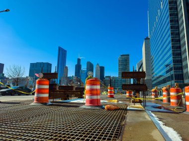 İşçiler Chicago şehir merkezinde yol inşaatı yapıyorlar. Turuncu trafik bariyerleri bölgeyi kapatırken, güneşli bir öğleden sonra arka planda yüksek ofis binaları yükseliyor..