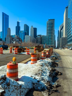 Chicago 'da turuncu trafik konilerinin bölgeyi işaretlediği bir yol çalışması yapılıyor. Kar hala yerde görülebilir durumda. Uzun binalar açık mavi gökyüzüne karşı arkaplan oluşturur.