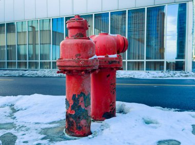 İki kırmızı yangın musluğu modern bir binanın yanına yerleştirilir. Etraflarını kar kaplıyor. Cam pencereler arka planda şehrin ufuk çizgisini yansıtıyor.