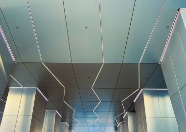 Manzara modern bir binanın tavanına bakıyor. Tasarım, parlak ışığı yansıtan metalik yüzeyli zikzak desenleri içeriyor. Gün ortası ayarı..