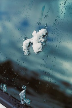 Yağmur damlaları kayarken kar bir arabanın camında durur. Gökyüzü bulutlu ve gri, bu da kış havasını gösteriyor. Bu sahne soğuk bir öğleden sonrayı gösteriyor..