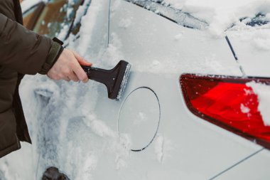 Bir kişi karla kaplı bir arabanın yanında duruyor ve sabah vakti aracın arkasındaki karı temizlemek için bir kazıyıcı kullanıyor..