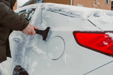 Birisi binanın dışında park etmiş bir arabadan kar çıkarmak için kazıyıcı kullanıyor. Kış mevsimindeyiz ve kar aracı ve çevreyi kaplıyor. Gün ışığı görünür..
