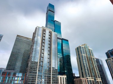 Yoğun bir şehirde gökdelenler bulutlu bir gökyüzüne karşı dik dururlar. Yansımalar cam pencerelerden yansıyor. İnsanlar aşağıdaki sokaklara taşınıyor, şehir yaşamını ve modern tasarımı vurguluyorlar..