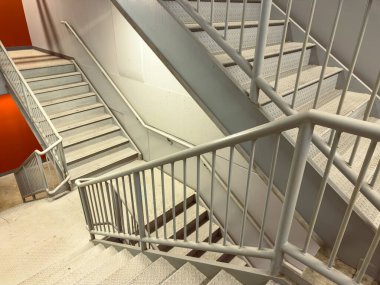 Binanın içindeki bir merdiven beyaz korkulukları ve farklı seviyelere çıkan basamakları gösteriyor. Bölge iyi ışıklandırılmış, turuncu duvarlarla çevrili. Bu da insanların kullanabileceği işlevsel bir alan anlamına geliyor..