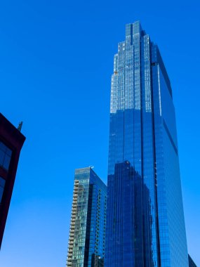 Uzun cam gökdelen açık mavi gökyüzünün altında şehir manzarasında duruyor. Güneş ışığı binanın pencerelerinden yansıyor. Yakın binalar farklı mimari tarzlar gösteriyor.