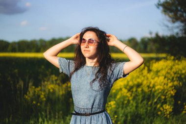 Sarı çiçeklerle dolu bir tarlada güneş gözlüğü ve kot bir elbise takan genç bir kadın ellerini saçlarına kaldırıyor ve mavi gökyüzünün altında rahatlamış görünüyor.