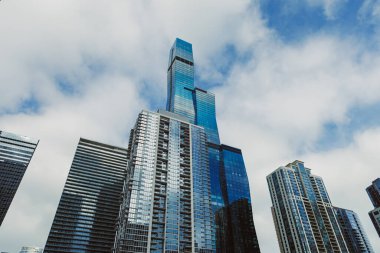 Yüksek binalar bulutlu bir gökyüzünün altında parlak bir günde camlardan yansıyan güneş ışığıyla bir şehir silüeti oluşturur.