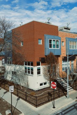 Sahne, şehir caddesindeki modern bir binayı gösteriyor. Üzerinde ağaçlar ve açık mavi bir gökyüzü bulunur. Yakınlarda sokak yönünü gösteren trafik işaretleri var..