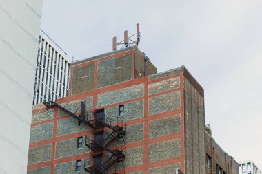 Tuğla bina, çatıda antenleri olan bir yangın çıkışını gösteriyor. Çevredeki binalar şehir manzarasına eklenir. Sahne, gün boyunca açık bir gökyüzünü yakalar..