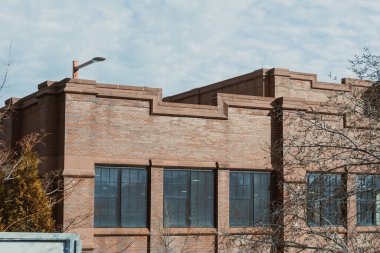 Manzara bulutlu bir gökyüzünün altında uzun pencereli tuğla bir bina gösteriyor. Kentsel bir ortam ve ortak bir mimari tarzı temsil ediyor..