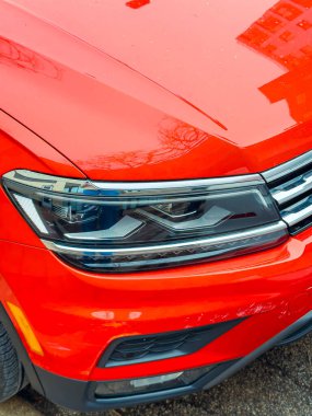 Kırmızı bir araba arka planda binalar olan bir caddede park halinde. Odak noktası arabanın ön tamponu ve farları. Sahne gündüzleri çekiliyor..