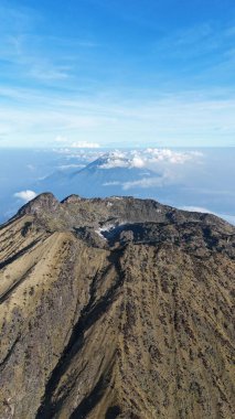 Yumuşak bulutlarla benekli açık mavi gökyüzüne karşı bir dağ sırasının havadan görünüşü. Engebeli arazi, seyrek bitki örtüsünden çorak kayaya dönüşerek güneş ışığı altında çarpıcı bir kontrastlık yaratır..