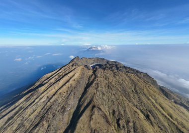 Büyüleyici bir hava görüntüsü bulutlar denizinin üzerindeki dingin, mavi gökyüzüne uzanan bir dağ zirvesini gözler önüne seriyor. Dağın yamaçları seyrek bitki örtüsüyle kaplı, engebeli arazisini ve doğal güzelliğini vurguluyor..