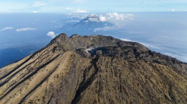 Bu, engebeli arazisini ve kraterini gösteren bir volkanın havadan çekilmiş görüntüsü. Uzakta, bulutların üzerinde başka bir yanardağ yükselir ve manzaraya derinlik katar. Gökyüzü açık, bu çekim için mükemmel..