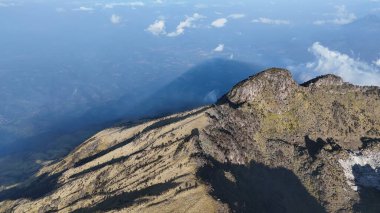Yüksek açılı bir manzara, bir dağ sırtının engebeli güzelliğini, güneş ışığıyla yıkanmış çimenli yamaçlarını yakalar. Gökyüzü açık mavi, dağların gölgeleriyle işaretlenmiş..