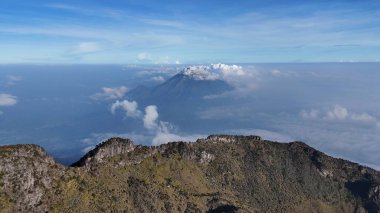 Yüksek açılı bir manzara bulutlarla kaplı engebeli bir dağ sırasını yakalar. Gökyüzü açık mavi, dağ manzarasının doğal güzelliğini ve huzurunu arttırıyor..