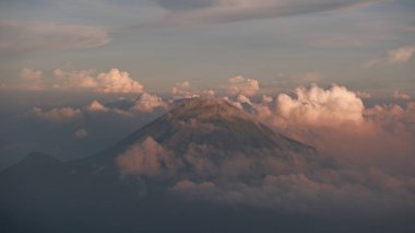 Gün batımında tüylü beyaz bulutlar tarafından gizlenmiş yüksek bir dağın çarpıcı manzarası. Gün batımından gelen yumuşak ışık bulutları hassas bir pastel tonla boyuyor, davetkar ve sakin bir atmosfer yaratıyor..