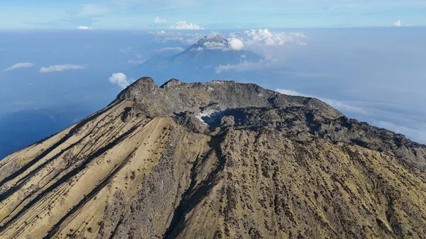 Yukarıdan yakalanan geniş bir sıradağ, bulutlarla kaplı bir gökyüzüne karşı iki belirgin tepenin yer aldığı araziye doğru uzanıyor. Öndeki dağ kurak bitki örtüsüne ve engebeli araziye sahiptir..