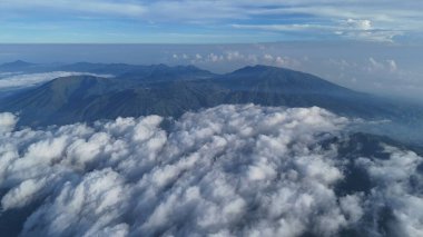 Yüksek açılı çekimde yoğun ve yuvarlanan bulutların arasından manzaralı bir dağ sırası görünüyor. Seyahat, doğa, macera ya da huzur temalı projeler için ideal. Hayret ve saygı uyandırır.