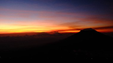 Canlı ve sıcak bir günbatımına karşı karanlık bir dağ siluetinin yer aldığı çarpıcı bir manzara. Seyahat, doğa ya da macera teması için mükemmel. Huzuru ve korkuyu çağrıştırıyor