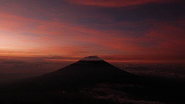 Dramatik manzara, canlı bir günbatımında siluetli bir volkanı yansıtıyor. Kırmızı ve turuncu renkler gökyüzünü boyuyor, huzur hissi uyandırıyor. Seyahat, doğa, macera temaları için ideal