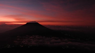 Görkemli bir volkan silueti şafak vakti bulutları delip geçer. Kızıl gökyüzü dram ve gizemi çağrıştırıyor. Seyahat, macera ya da doğa teması için mükemmel. Atmosferik. Nefes kesici.