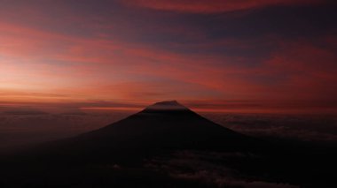 Dramatik manzara, canlı bir günbatımında siluetli bir volkanı yansıtıyor. Kırmızı ve turuncu renkler gökyüzünü boyuyor, huzur hissi uyandırıyor. Seyahat, doğa, macera temaları için ideal