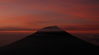 Renkli bir günbatımı gökyüzüne karşı bir volkanın karanlık silueti. Sıcak renkler huzurlu ve dramatik bir sahne yaratır. Doğa, seyahat ya da ilham kullanımı için ideal. Zirvenin altındaki bulut tabakası