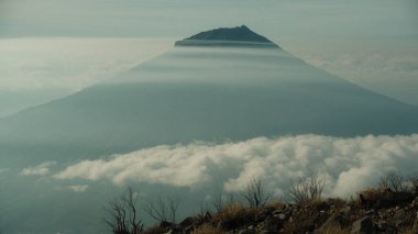 Görkemli bir dağ zirvesi yoğun, kabarık bulutların üzerinde yükselir. Sis arazide yuvarlanıyor. Sakin, huzurlu bir atmosfer. Doğa, seyahat ve sükunet temaları için ideal