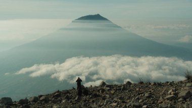 Bir dağ zirvesinin tepesinde yürüyüş yapan ve bulutların ortasındaki görkemli bir volkanın tadını çıkaran hayranlık uyandırıcı bir manzara. Ruh hali sakin, macera, seyahat ve doğa teması için ideal.