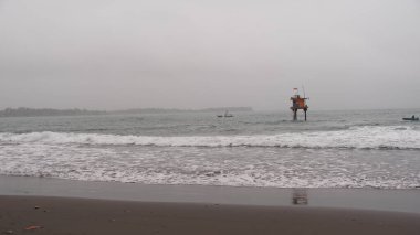 Sisli bir plajın sükunetini bu görüntüyle yakalayın. Denizde bir kule ve uzak tekneler gösteriyor, seyahat için mükemmel, sahil temaları, doğa terfisi, ve barışçıl ruh hali. Web siteleri, broşürler ve reklam kullanımı için ideal