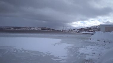Karla kaplı bir arazi, küçük bir kıyı yerleşimi ve solgun bir gökyüzünün altında engebeli dağların yer aldığı sakin bir kış manzarası. Soğuk iklim, mevsimsel hava ve huzurlu kırsal çevre.