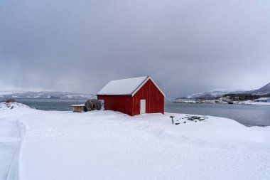 Karla kaplı yolu denize, geleneksel evleri ve açık mavi gökyüzünün altındaki dağları çevreleyen küçük bir kıyı köyünün kış manzarası. Soğuk iklim ve kırsal mimari ile barışçıl İskandinav manzarası.