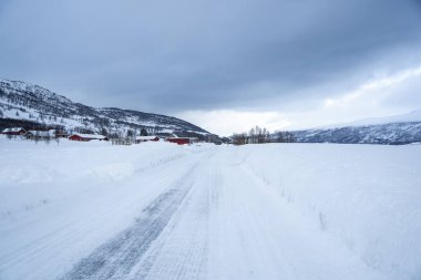 Karla kaplı yolu denize, geleneksel evleri ve açık mavi gökyüzünün altındaki dağları çevreleyen küçük bir kıyı köyünün kış manzarası. Soğuk iklim ve kırsal mimari ile barışçıl İskandinav manzarası.