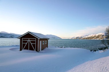 Karla kaplı yolu denize, geleneksel evleri ve açık mavi gökyüzünün altındaki dağları çevreleyen küçük bir kıyı köyünün kış manzarası. Soğuk iklim ve kırsal mimari ile barışçıl İskandinav manzarası.
