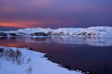 Karla kaplı yolu denize, geleneksel evleri ve açık mavi gökyüzünün altındaki dağları çevreleyen küçük bir kıyı köyünün kış manzarası. Soğuk iklim ve kırsal mimari ile barışçıl İskandinav manzarası.