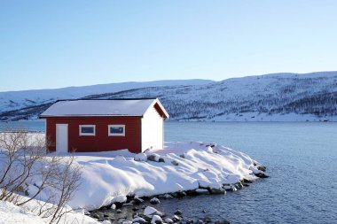 Karla kaplı yolu denize, geleneksel evleri ve açık mavi gökyüzünün altındaki dağları çevreleyen küçük bir kıyı köyünün kış manzarası. Soğuk iklim ve kırsal mimari ile barışçıl İskandinav manzarası.