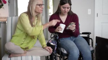 In a cozy living room, a daughter in a wheelchair connects with her mature mother, sharing happy memories through photos on a smartphone, enjoying the moment together.