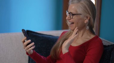 Uzun saçlı yaşlı bir kadın akıllı telefonuyla video sohbeti yaparken ışıl ışıl gülümsüyor, arka planda mavi bir duvarla koltukta rahatça oturuyor, neşeli bir anın tadını çıkarıyor..