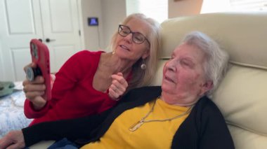 Yaşlı bir kadın, sağlık merkezindeki mobil bir cihazda ona bir şey gösteren bakıcısıyla ilgilenmekten zevk alır..