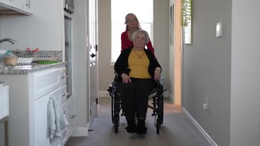 Tekerlekli sandalyedeki yaşlı kadın, ev sağlığı merkezinden geçerken bakıcıdan yardım alıyor.